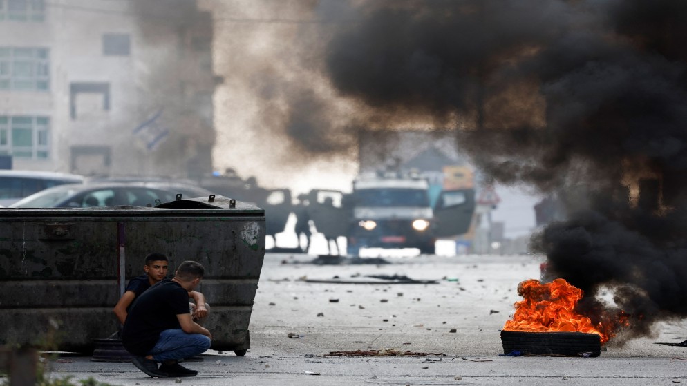 جنود الاحتلال الإسرائيلي يقفون بجوار مركبات عسكرية بينما يحتمي فلسطينيون بالقرب من إطار مشتعل في أثناء مداهمة إسرائيلية في قلنديا بالضفة الغربية المحتلة، 7 تشرين الأوّل/ أكتوبر 2024. (رويترز)